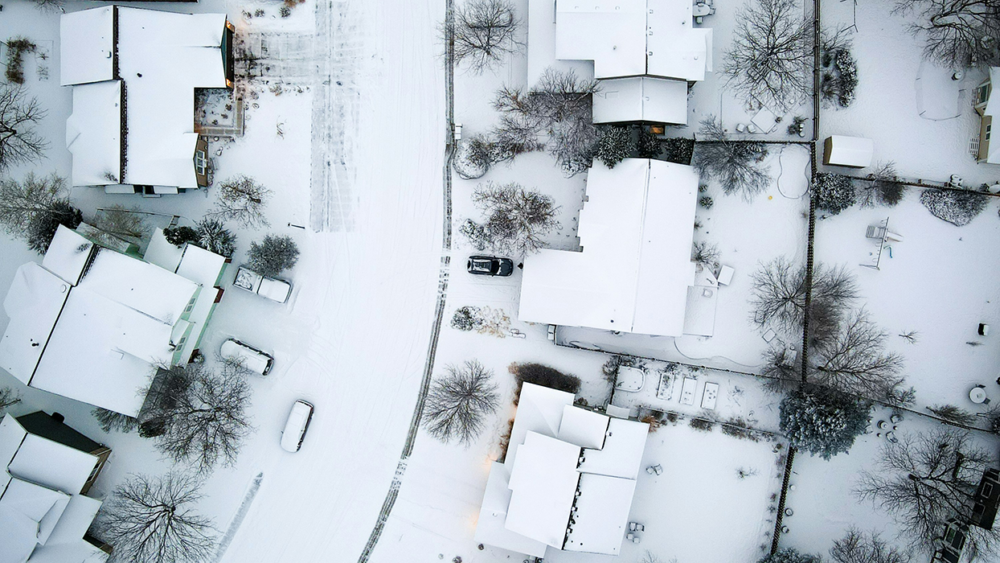 Aerial view of snow-covered suburban neighborhood with houses, trees, and parked cars. Roads and roofs are blanketed in snow, creating a wintery scene.